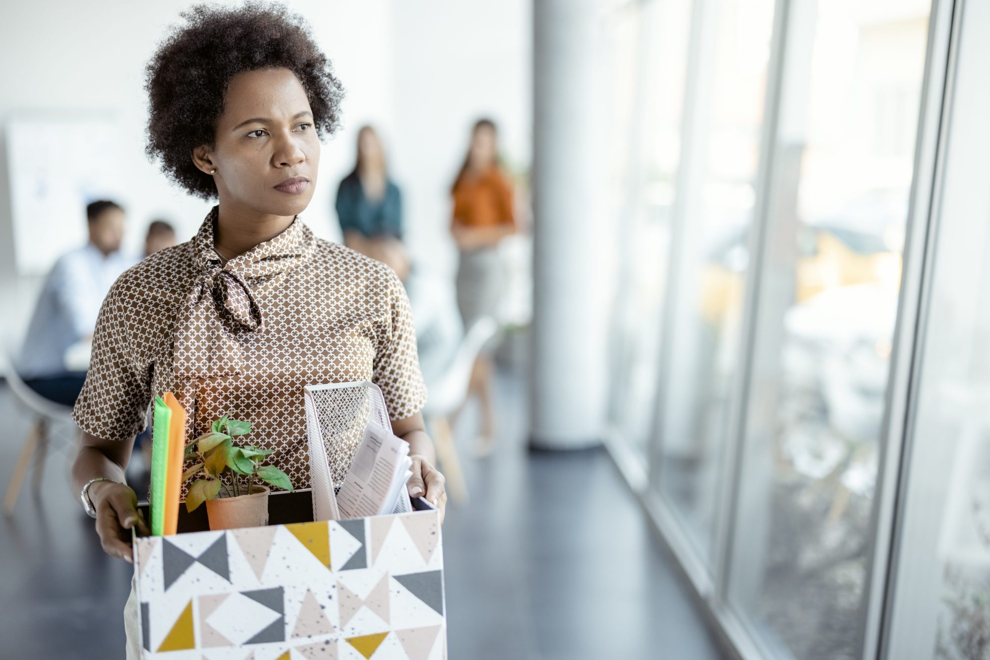 Mature african american woman resigning from work. Female walks through the office, carrying box with personal belongings. Business, resignation and job loss concept