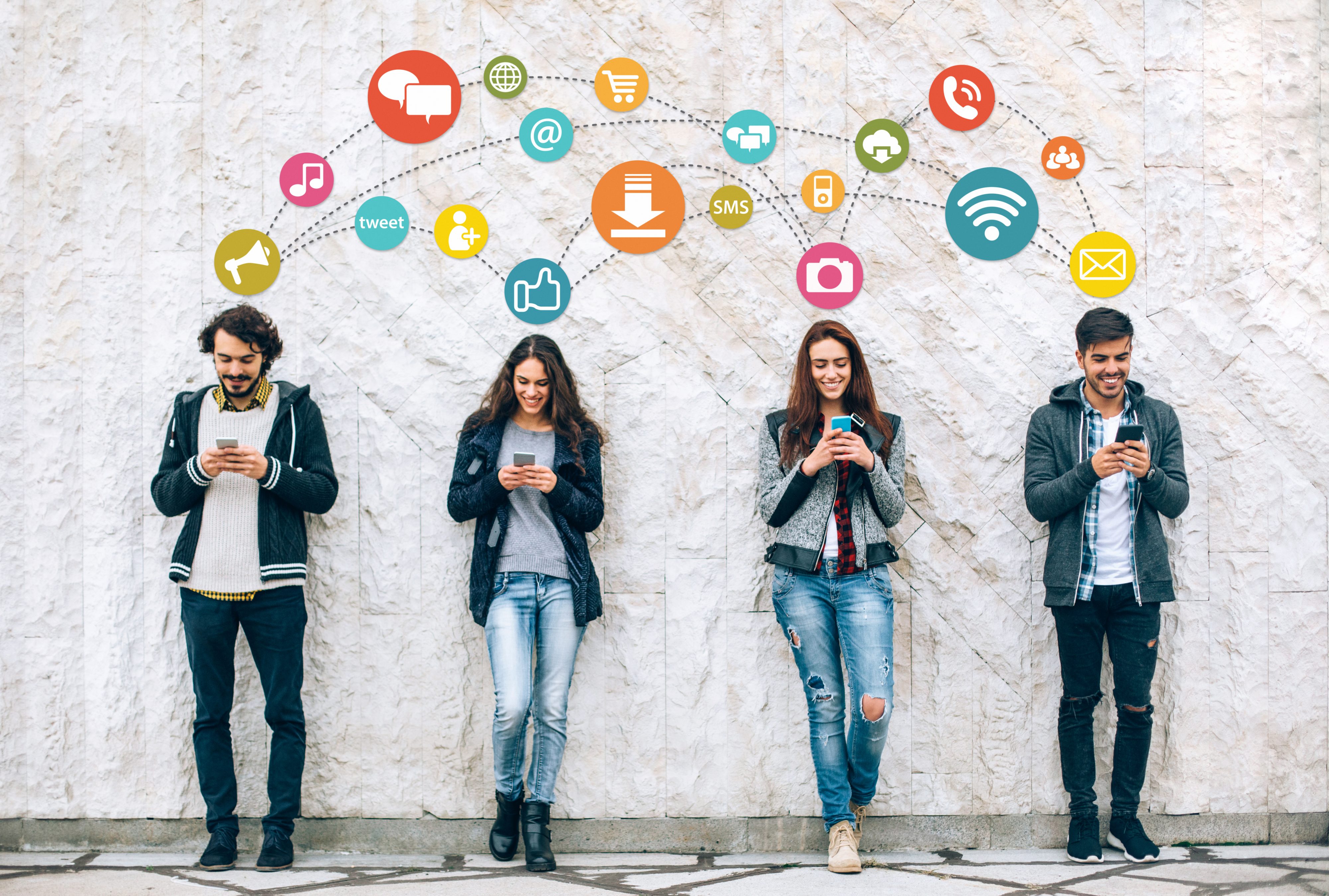 Social network concept with smiling young people holding smart phones in front of big white wall.