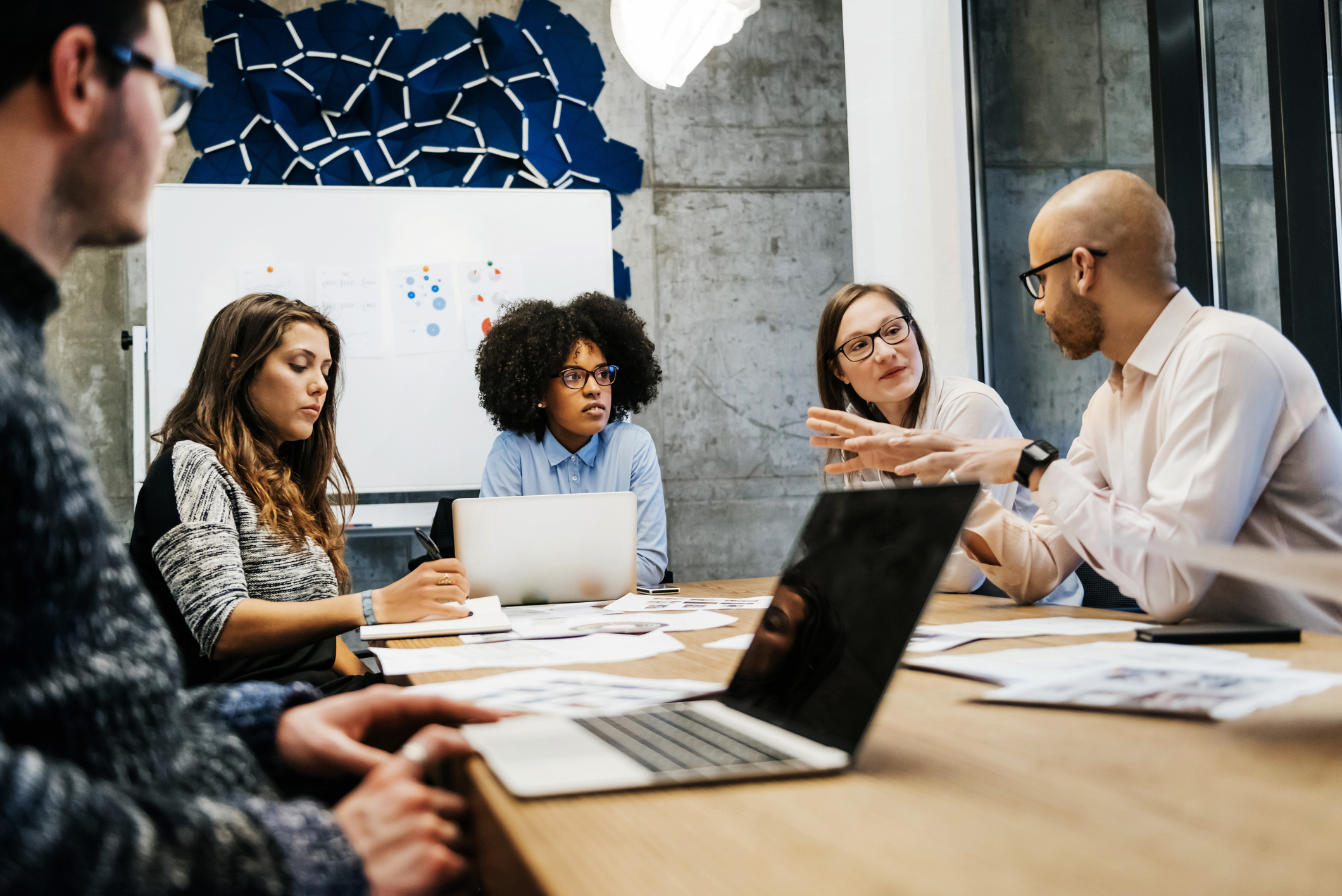 A group of three young women and two men of different ethnicities are in a business meeting in a modern day office. A bald man is talking to the group while there are laptops and documents on the table. They are discussing major gifts.