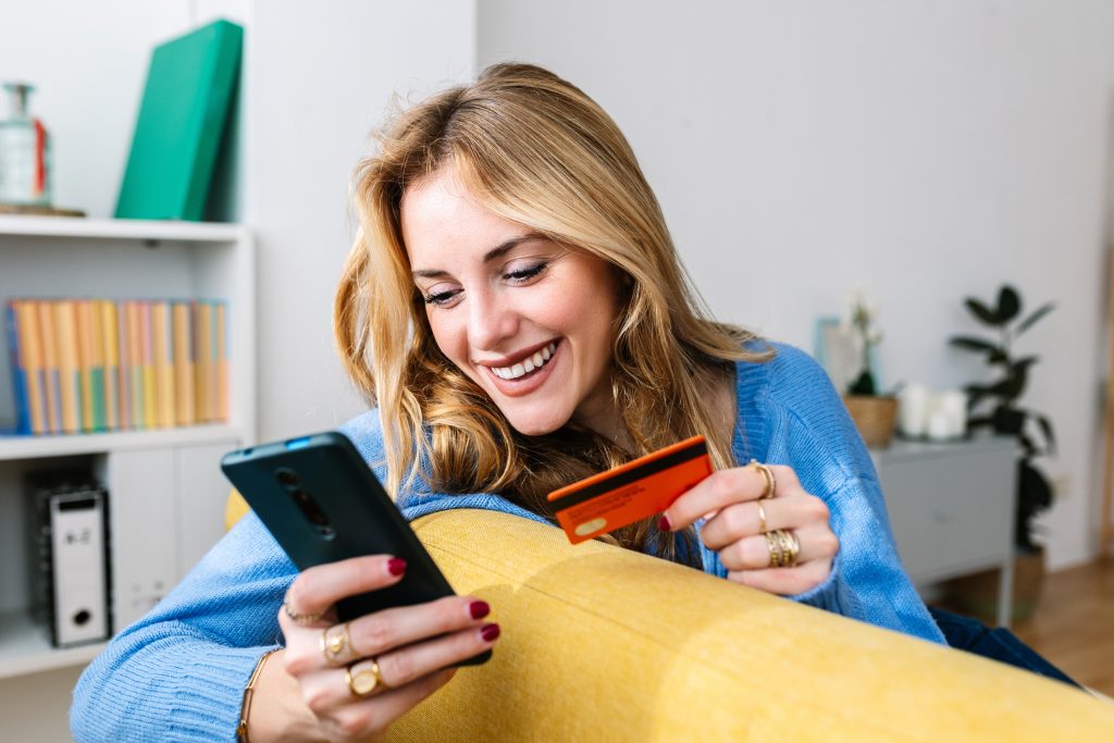 Happy young adult blonde woman holding credit card while making a donation using mobile phone app at home. E-commerce and delivery service concept.