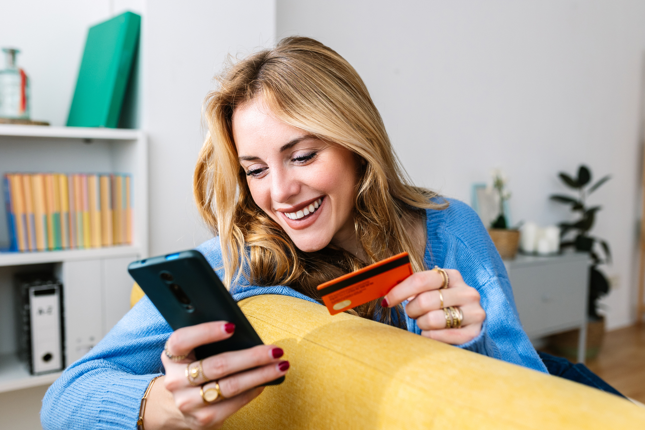 Happy young adult blonde woman holding credit card while making a donation using mobile phone app at home. E-commerce and delivery service concept.